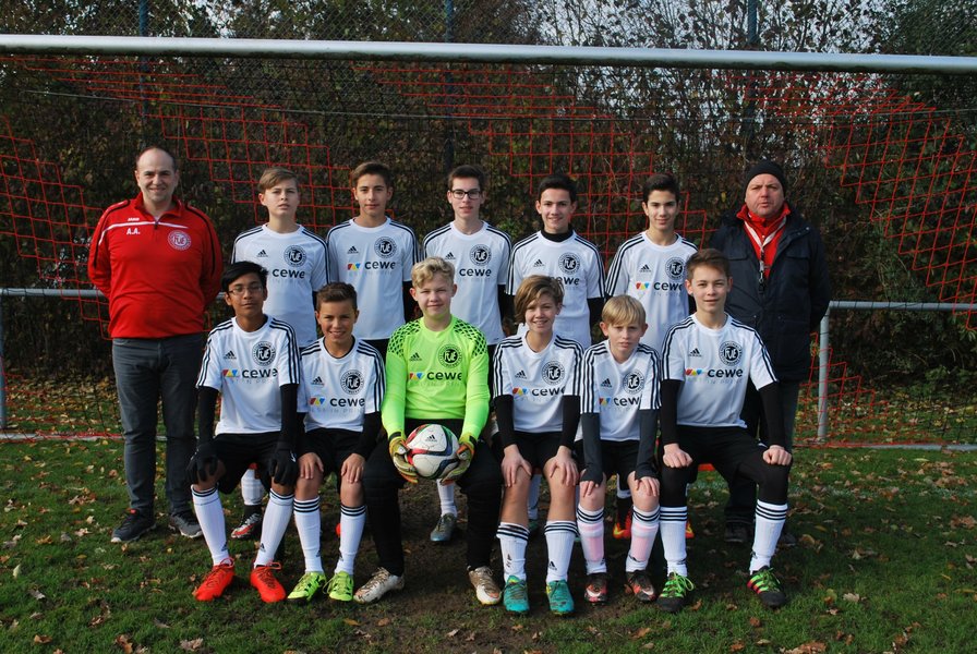 Eine Jugendfußballmannschaft posiert für ein Foto auf dem Feld. Sie tragen schwarz-weiße Uniformen. Auf beiden Seiten stehen zwei Trainer. Der Torwart im grünen Trikot hält einen Ball. Im Hintergrund sind Bäume und ein Fußballtor zu sehen.