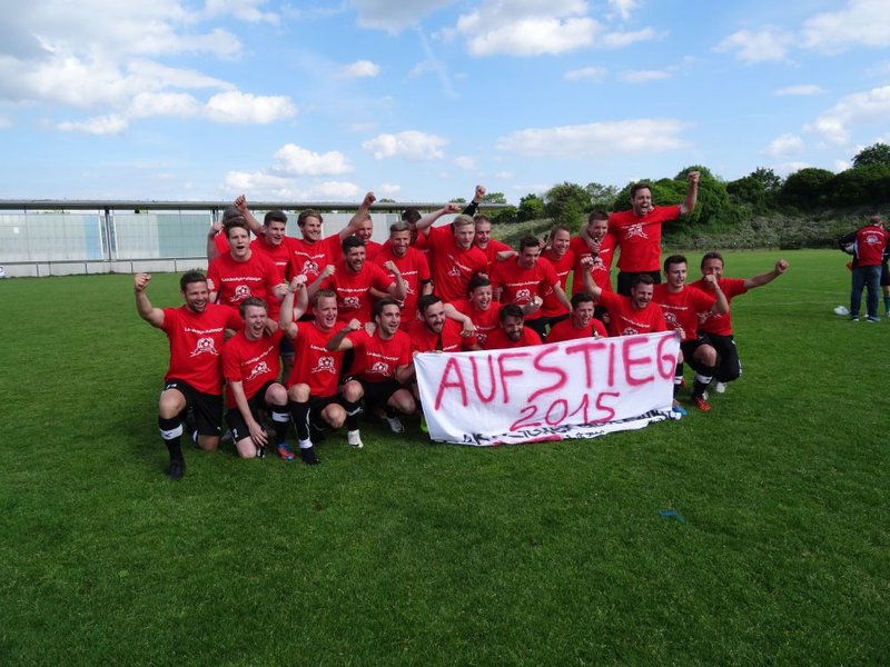 Eine Gruppe von Fußballspielern in roten Trikots feiert auf einer grünen Wiese und hält begeistert ein Banner mit der Aufschrift „AUFSTIEG 2015“ hoch.