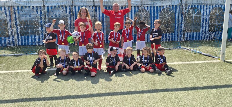 Eine Gruppe kleiner Kinder in Fußballtrikots posiert für ein Mannschaftsfoto auf einem Fußballfeld. Sie werden von zwei Erwachsenen begleitet. Einige Kinder halten Medaillen und eines hält einen Fußball. Im Hintergrund sind ein Metallzaun und ein Netz zu sehen.