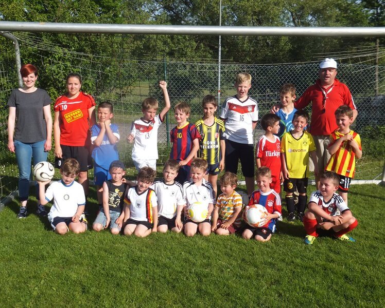 Eine Gruppe kleiner Kinder, einige davon in Fußballtrikots, posiert mit zwei Erwachsenen vor einem Fußballtor. Sie stehen an einem sonnigen Tag auf einer Wiese. Ein Kind hält einen Fußball und mehrere lächeln.