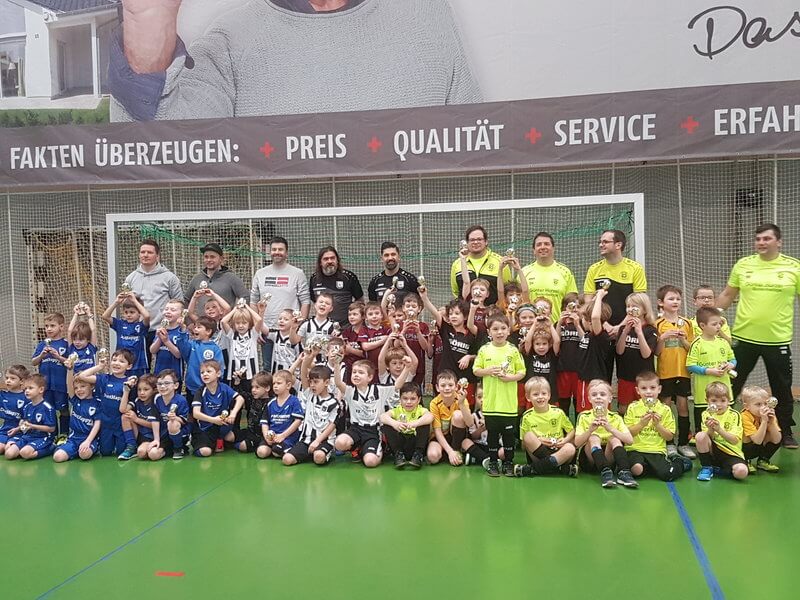 Eine Gruppe kleiner Kinder in verschiedenfarbigen Fußballtrikots posiert mit mehreren Erwachsenen in Sportkleidung in einer Turnhalle. Sie stehen vor einem Fußballtor und über ihnen ist teilweise ein großes Werbebanner zu sehen.