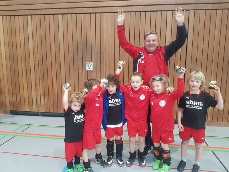 Eine Gruppe Kinder in roter Sportkleidung posiert mit einem Mann, ebenfalls in Sportkleidung. Sie befinden sich in einer Turnhalle mit Holzwänden. Die Kinder halten kleine Trophäen und lächeln, während der Mann seine Arme zum Jubeln streckt.