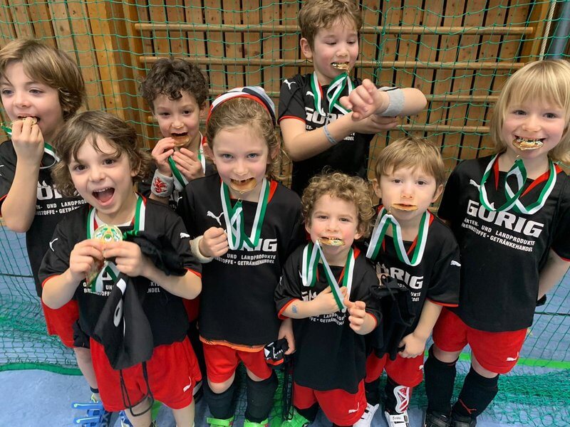 Eine Gruppe von Kindern steht in einer Sportumgebung. Sie tragen alle passende schwarz-rote Sportuniformen und halten Medaillen in den Händen. Sie tun spielerisch so, als würden sie in ihre Medaillen beißen und lächeln, um Aufregung und Freude auszudrücken. Im Hintergrund ist eine Netzwand zu sehen.