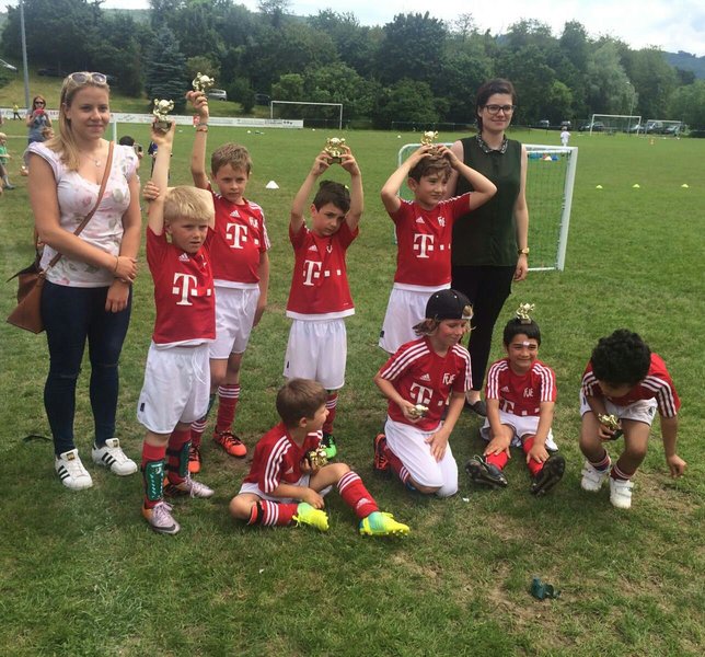 Eine Gruppe junger Jungen in passenden Fußballtrikots hält stolz ihre Trophäen hoch, während sie auf einem Rasen knien und stehen. Zwei Erwachsene stehen lächelnd hinter ihnen. Im Hintergrund sind Fußballtore und Bäume zu sehen.