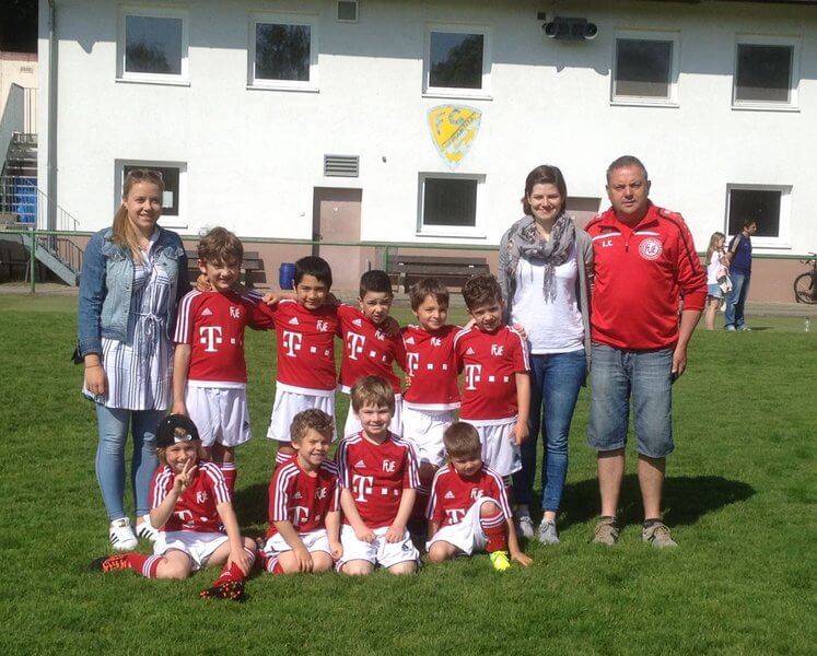 Eine Gruppe kleiner Kinder in rot-weißen Fußballtrikots posiert auf einer Wiese. Sie werden von zwei Frauen und einem Mann in Freizeitkleidung flankiert. Im Hintergrund ist ein Gebäude mit Fenstern und einem Logo zu sehen.