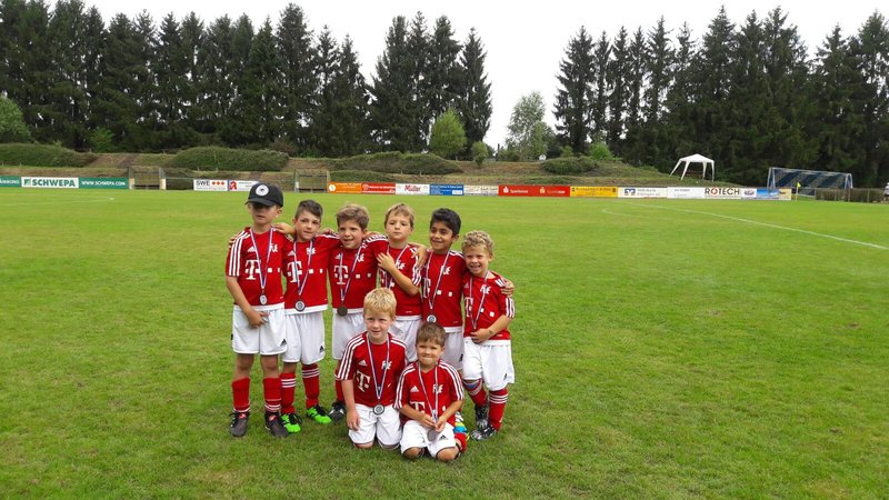 Eine Gruppe kleiner Kinder in rot-weißen Fußballtrikots steht zusammen auf einem Rasenplatz, jeder trägt eine Medaille. Im Hintergrund sind Bäume und Werbetafeln zu sehen. Sie wirken fröhlich und posieren für ein Mannschaftsfoto.