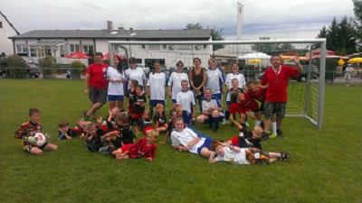 Eine Gruppe von Kindern und Erwachsenen posiert gemeinsam auf einem Fußballfeld, mit Torpfosten im Hintergrund, und feiert ihre Teamarbeit und ihren Sportsgeist.