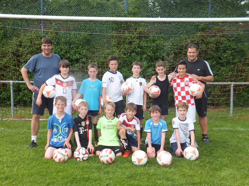 Eine Gruppe von jungen Fußballspielern und Trainern posiert gemeinsam vor einem Tor, hält Fußbälle und trägt verschiedene Mannschaftstrikots.