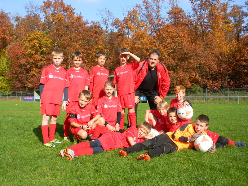 Eine Jugendfußballmannschaft in roten Trikots posiert auf einem von bunten Herbstbäumen umgebenen Rasenplatz. Der Trainer kniet zwischen den Spielern.