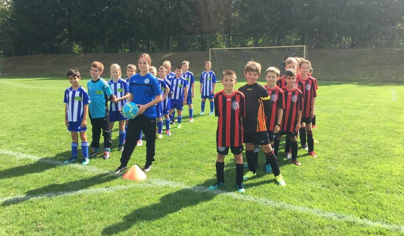 Zwei Kindermannschaften stehen auf einem Fußballplatz, eine in blau-weiß gestreiften Trikots, die andere in rot-schwarzen.