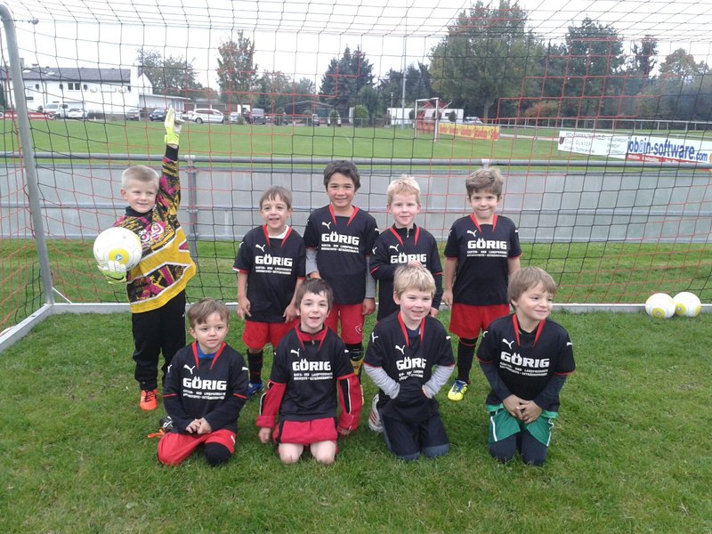 Eine Gruppe von neun kleinen Kindern in schwarzen Fußballtrikots posiert vor einem Tor. Einige halten Fußbälle in den Händen. Sie lächeln. Im Hintergrund sind eine Wiese und eine Straße mit Bäumen und Gebäuden zu sehen.