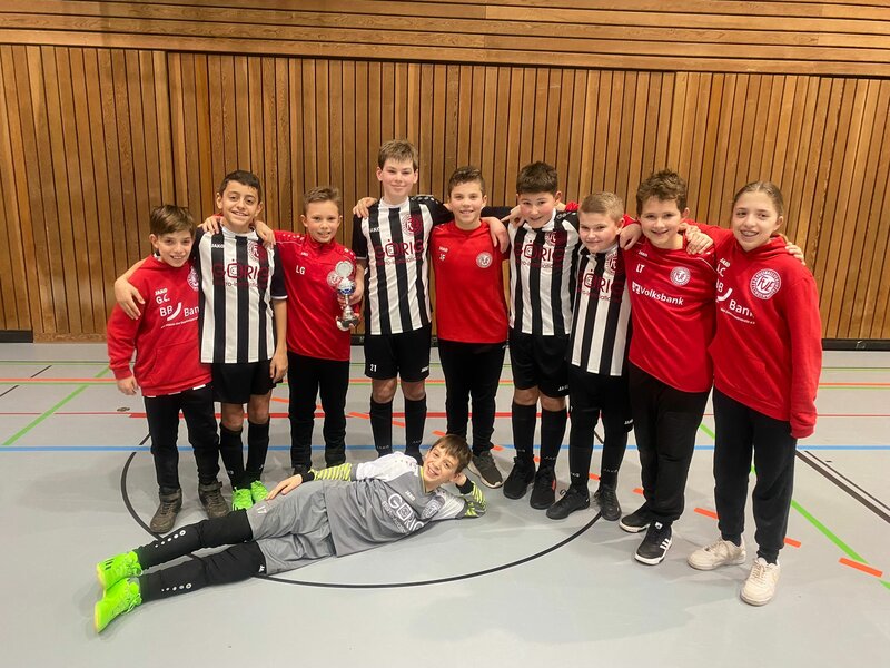Eine Jugendfußballmannschaft posiert in einer Turnhalle, trägt schwarz-weiß gestreifte Trikots und rote Sweatshirts, ein Mitglied hält einen Pokal in der Hand.