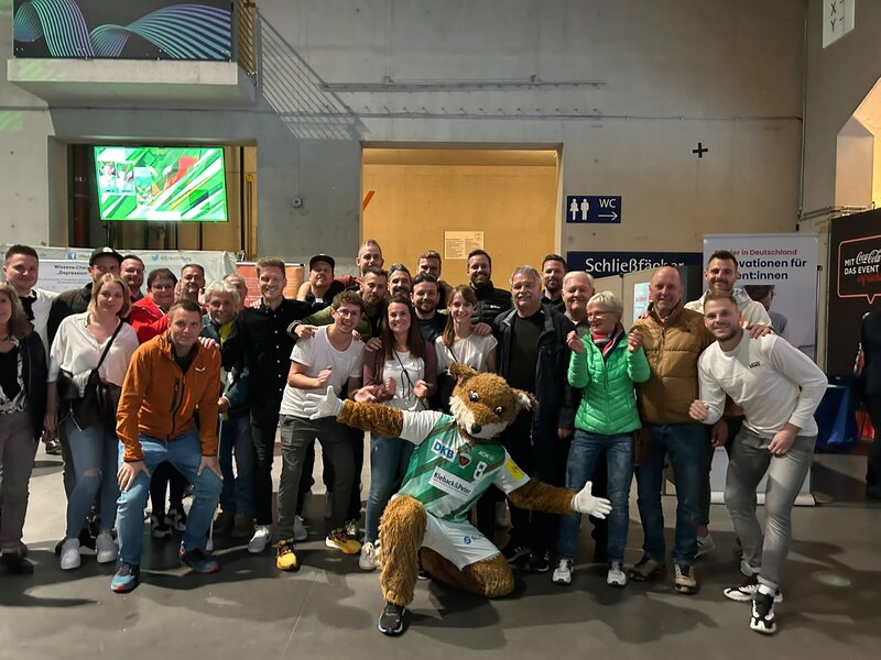 Eine Gruppe von Personen lächelt und posiert mit einem Maskottchen in einem Innenraum.