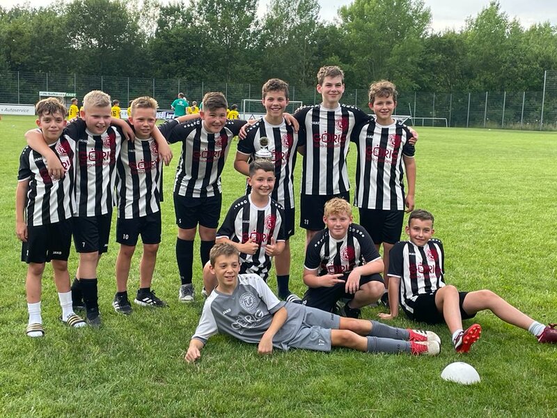 Eine Gruppe junger Fußballspieler in schwarz-weißen Trikots posiert gemeinsam auf einer Wiese, einige haben einen Pokal dabei und lächeln.