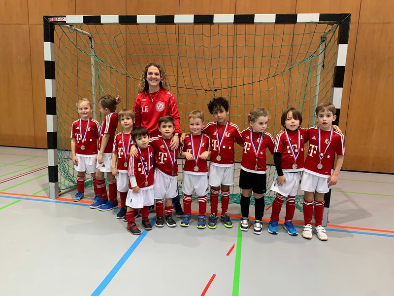 Eine junge Fußballmannschaft in roten Trikots posiert mit ihrem Trainer vor einem Tor in einer Halle. Jedes Kind trägt eine Medaille um den Hals.