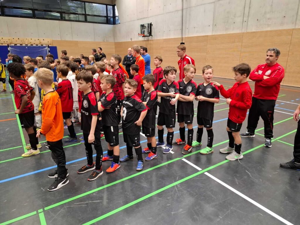 Eine Gruppe junger Fußballspieler in passenden schwarz-roten Trikots steht in einer Turnhalle und wartet auf Anweisungen, während die Trainer in der Nähe sind.