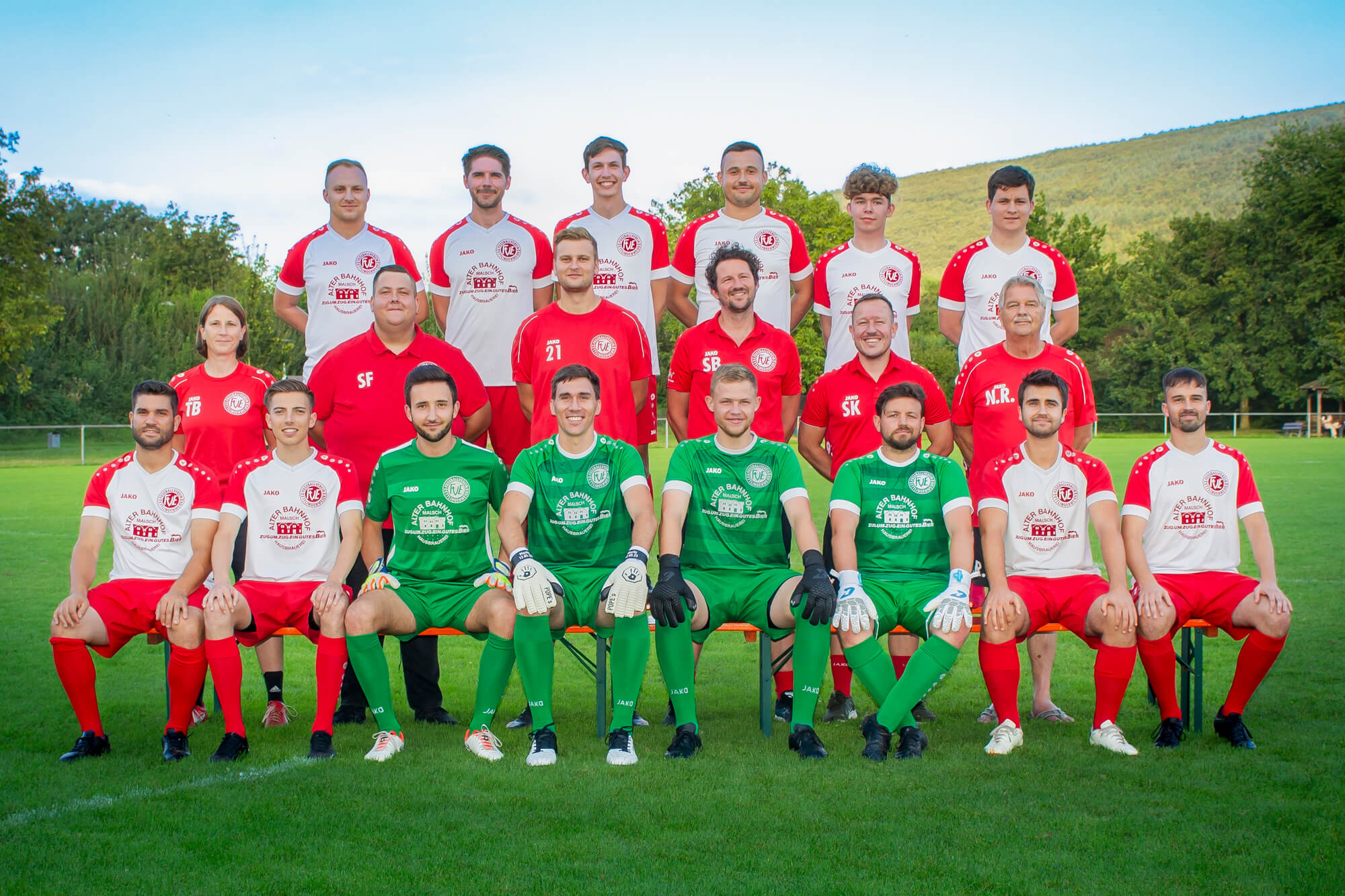 Ein Gruppenfoto einer Fußballmannschaft mit Spielern in roten und grünen Trikots, die im Freien auf einer Wiese mit Bäumen und einem Hügel im Hintergrund posieren.