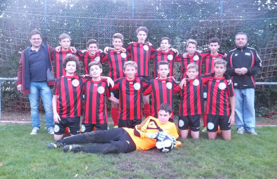 Eine Jugendfußballmannschaft posiert für ein Gruppenfoto auf einer Wiese. Die Spieler in schwarz-rot gestreiften Trikots stehen und knien vor einem Tornetz. Der Torwart im gelben Trikot hält einen Fußball vor sich liegend. Zwei Erwachsene stehen auf beiden Seiten.