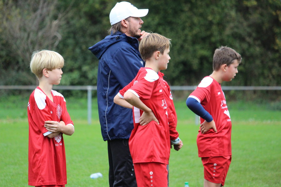 Eine Gruppe junger Fußballspieler in roten Trikots steht auf einem grünen Feld und beobachtet aufmerksam einen Trainer in der Nähe.