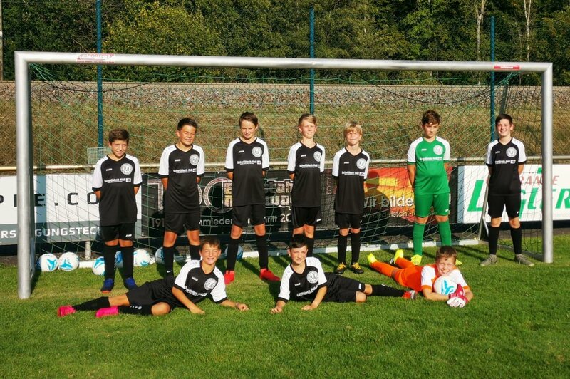Eine Jugendfußballmannschaft stellt sich für ein Gruppenfoto vor einem Tor auf. Sie trägt schwarz-weiße Trikots mit verschiedenen bunten Stutzen.