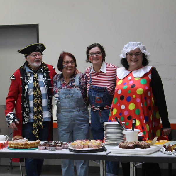 Eine Gruppe von vier Personen in farbenfrohen Kostümen steht neben einem Tisch, der mit verschiedenen Süßspeisen, darunter Kuchen und Gebäck, gefüllt ist.