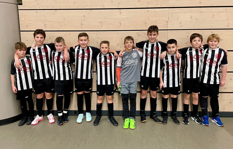 Eine Gruppe junger Fußballspieler in schwarz-weiß gestreiften Trikots steht gemeinsam vor einer Holzwand und lächelt in die Kamera.