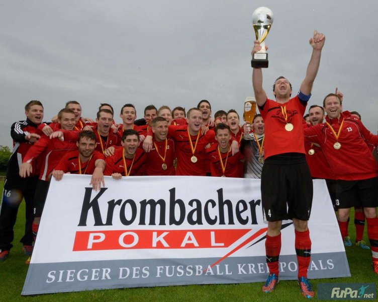 Eine jubelnde Fußballmannschaft in roten Trikots feiert mit einer Trophäe, die Goldmedaillen trägt, vor einem Krombacher Pokal-Banner.