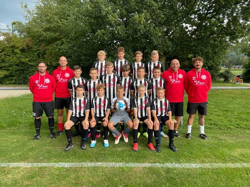 Eine Jugendfußballmannschaft posiert in schwarz-weiß gestreiften Trikots auf einer Wiese, während die Trainer in roten Hemden in der Nähe stehen.