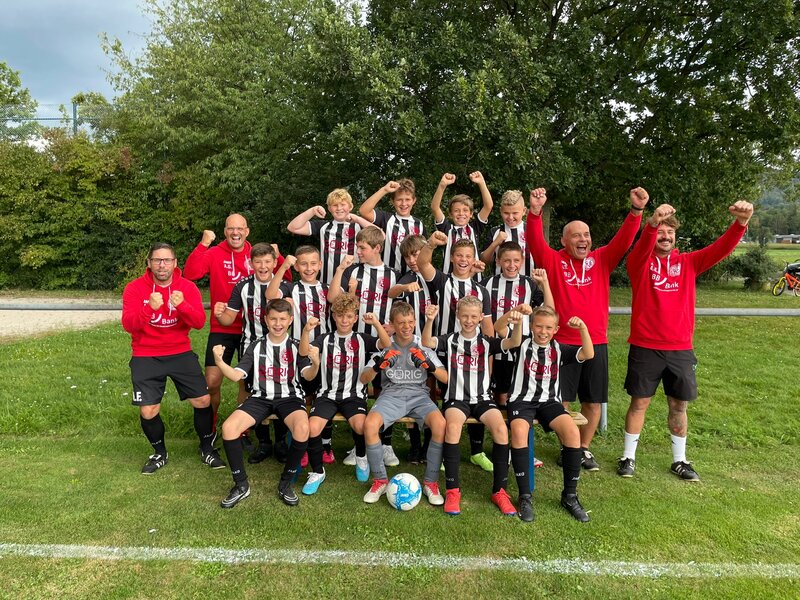 Eine Gruppe enthusiastischer junger Fußballspieler und -trainer in schwarz-weiß gestreiften Trikots feiert gemeinsam auf einer Wiese.