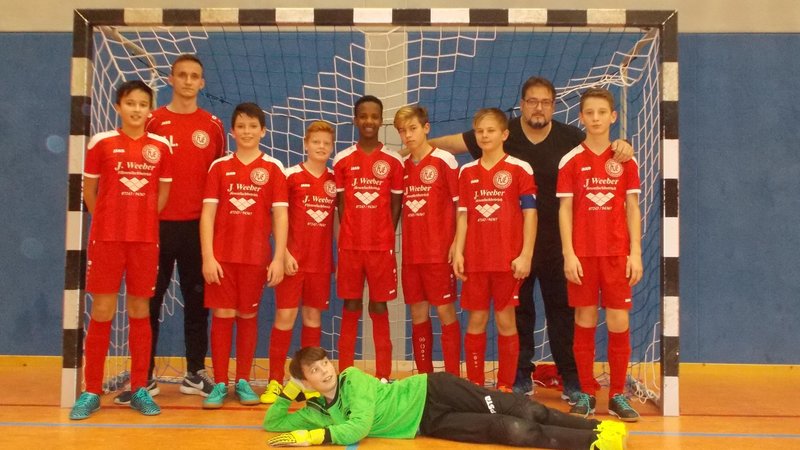 Eine Jugend-Hallenfußballmannschaft posiert in roten Trikots vor einem Tor, mit einem auf dem Boden liegenden Torwart und dahinter stehenden Mannschaftskameraden.
