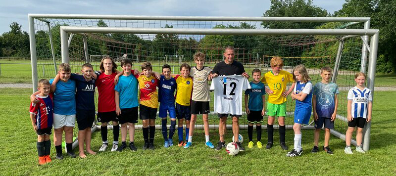 Eine Gruppe junger Fußballspieler und ein Trainer posieren gemeinsam vor einem Tor, halten ein Trikot und lächeln auf einer Wiese.