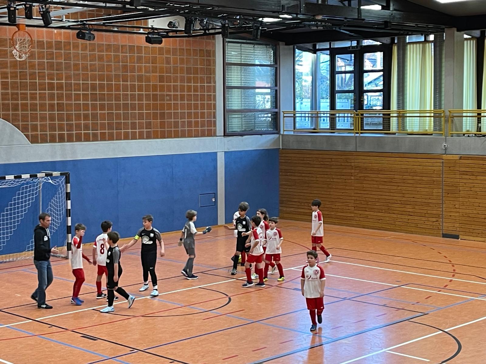 Junge Spieler in rot-weißen Trikots versammeln sich auf einem Hallenfußballplatz und feiern ein Tor oder den Abschluss eines Spiels.