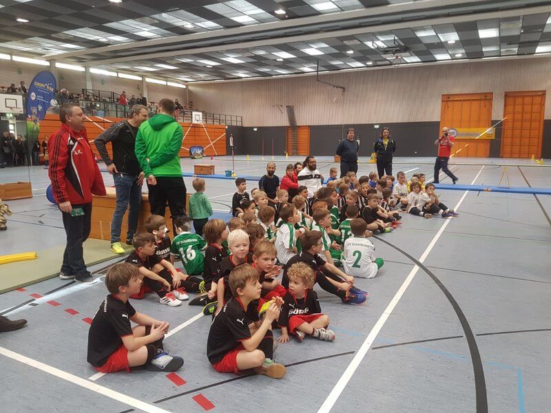 Eine Gruppe Kinder sitzt in verschiedenfarbiger Sportkleidung auf dem Boden einer Sporthalle. Um sie herum stehen Erwachsene in Freizeitkleidung. Die Halle ist geräumig, hat eine hohe Decke und im Hintergrund Trainingsgeräte.
