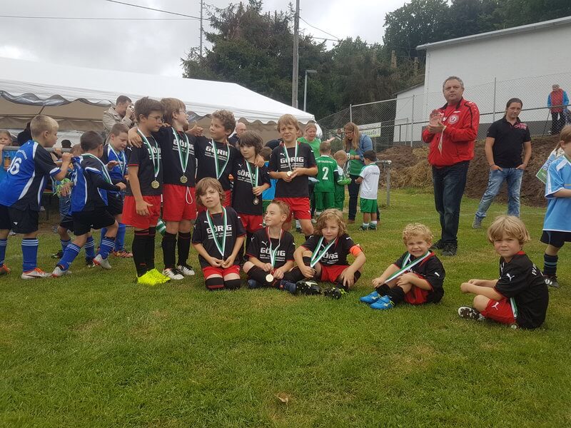 Eine Gruppe kleiner Kinder in Fußballuniformen und mit Medaillen posiert gemeinsam auf einer Wiese. Einige stehen, andere sitzen im Schneidersitz. Erwachsene und andere Kinder stehen im Hintergrund neben einem weißen Zelt. Bäume und ein Zaun sind zu sehen.