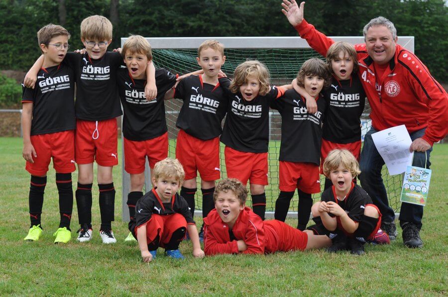 Eine Gruppe junger Jungen in schwarz-roten Fußballtrikots posiert fröhlich vor einem Tor auf einem Rasenplatz. Sie werden von einem Erwachsenen in einer roten Jacke begleitet, der lächelt und eine Hand hebt, in der anderen hält er ein Stück Papier.