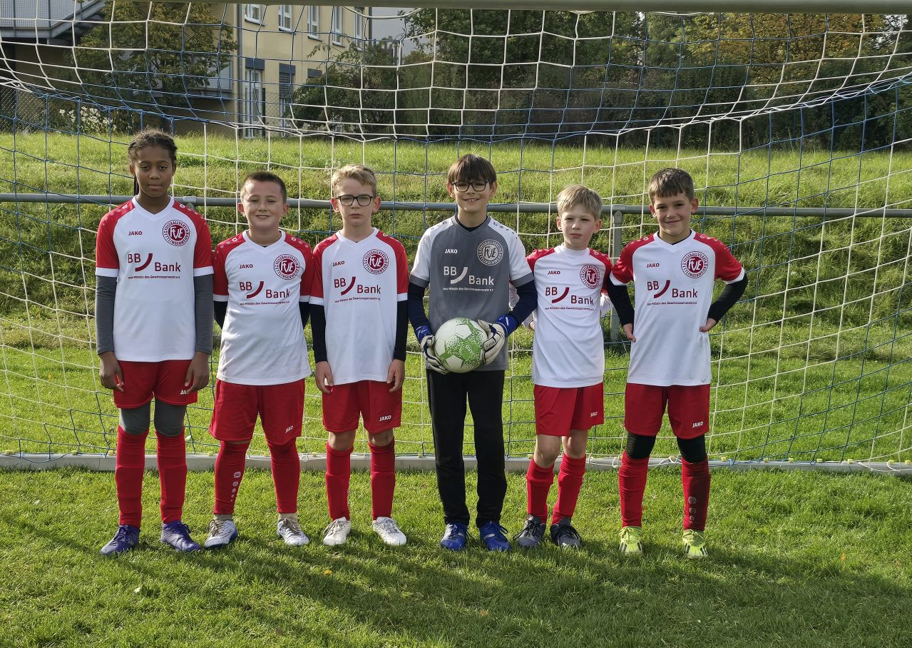 Eine Jugendfußballmannschaft posiert in rot-weißen Trikots vor einem Tor, wobei ein Spieler einen Fußball hält.