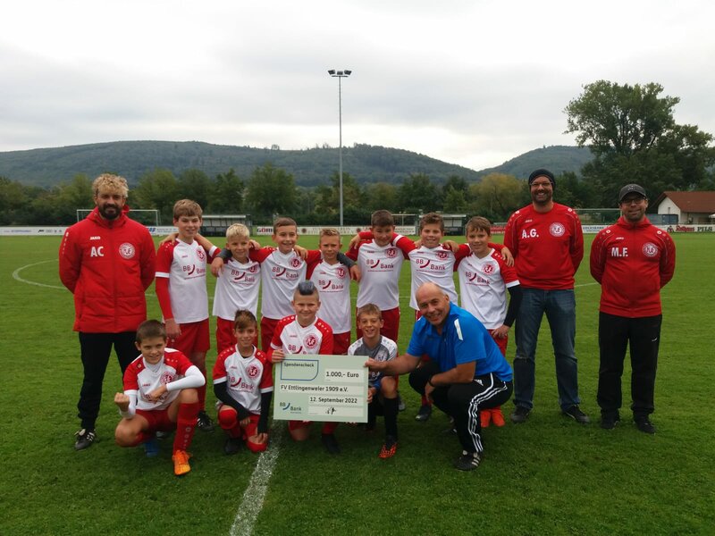 Eine Jugendfußballmannschaft in rot-weißen Trikots hält einen großen Scheck über 1.000 Euro in der Hand und feiert ihren Erfolg auf einem grünen Feld.