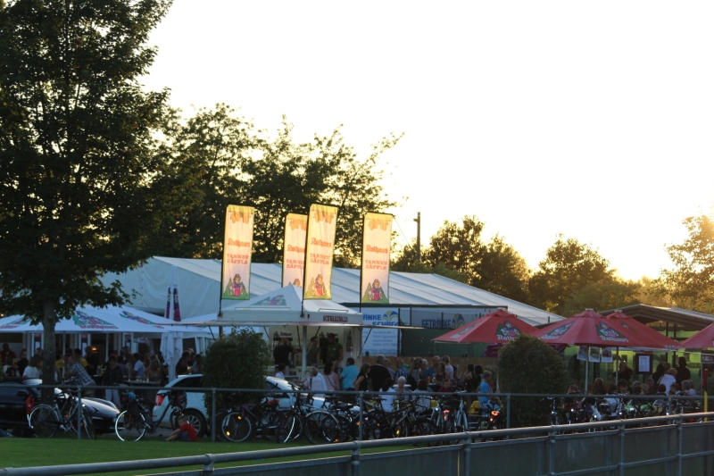 Eine lebhafte Szene im Freien bei Sonnenuntergang mit Zelten, Sonnenschirmen und Menschenmassen, die sich unter geparkte Fahrräder mischen.