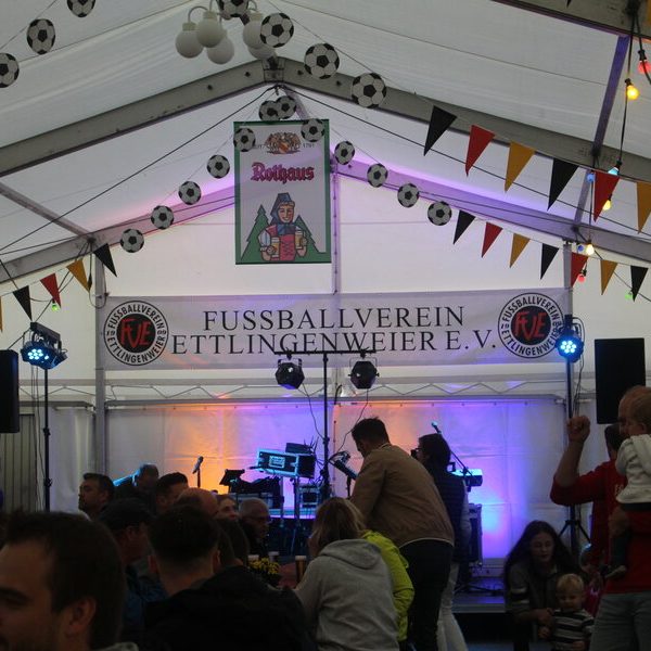 Ein festliches, mit Bannern zum Thema Fußball geschmücktes Zelt mit Bühne und Beleuchtung, gefüllt mit Menschen, die eine Veranstaltung genießen.