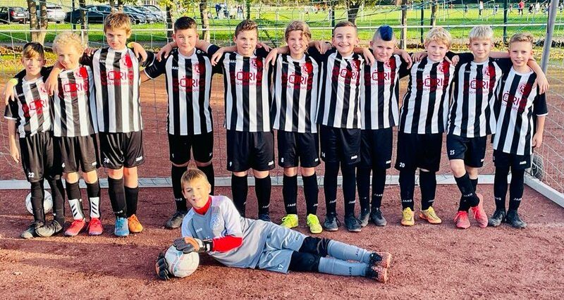 Eine Jugendfußballmannschaft posiert in schwarz-weiß gestreiften Trikots auf einem Feld. Der Torwart liegt vorne, bereit für den Einsatz.