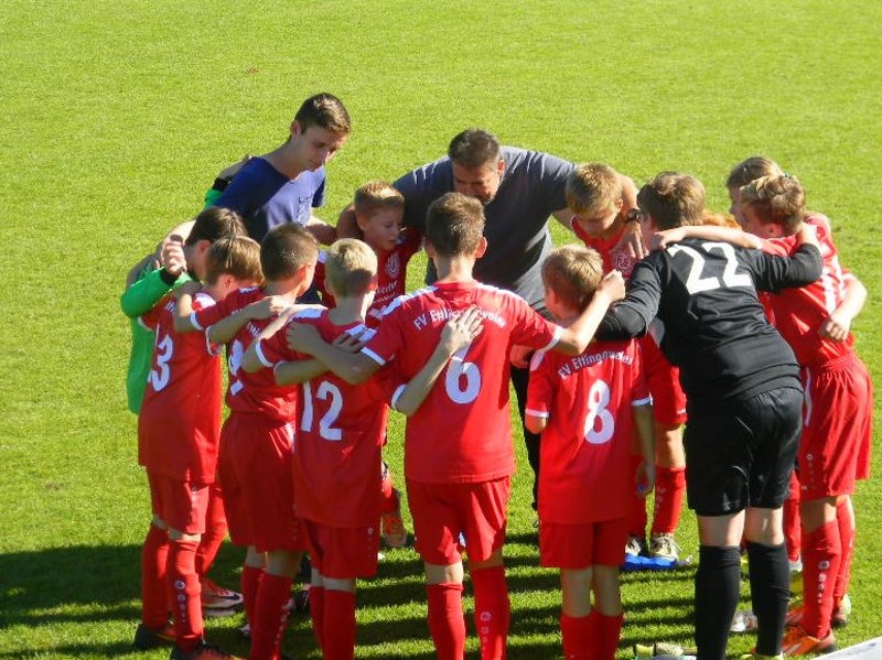 Eine Jugendfußballmannschaft in roten Trikots kauert auf einem sonnigen Feld zusammen und berät sich vor ihrem Spiel.