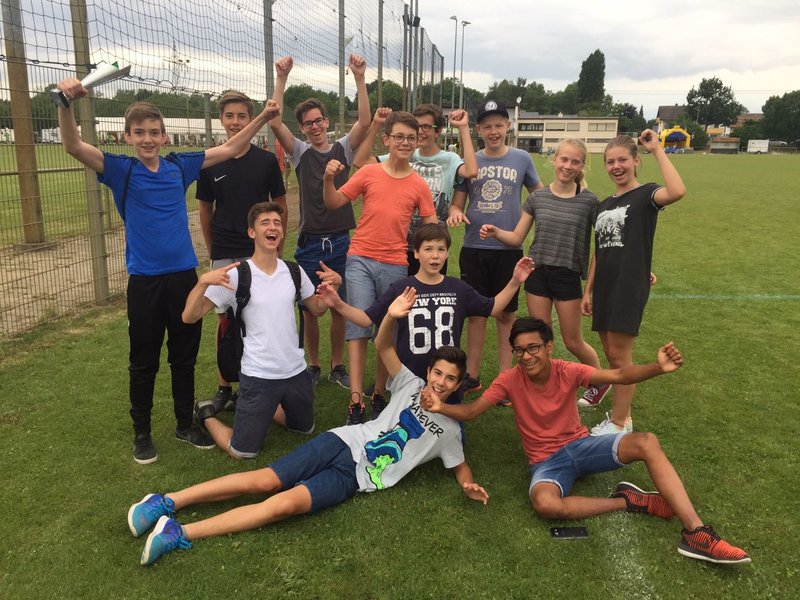 Eine Gruppe lächelnder Teenager posiert voller Energie auf einer Wiese, einige stehen, andere sitzen. Sie sind leger gekleidet und heben ihre Arme zum Feiern oder Jubeln. Im Hintergrund sind ein Zaun, Gebäude und Bäume zu sehen.