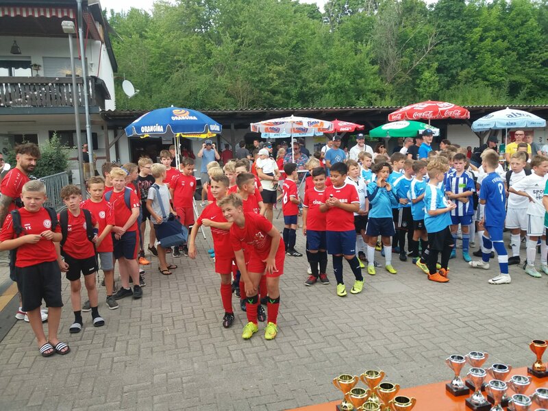 Eine Gruppe junger Fußballspieler in verschiedenen Mannschaftstrikots versammelt sich um einen Tisch mit Trophäen vor einem baumbestandenen Hintergrund.