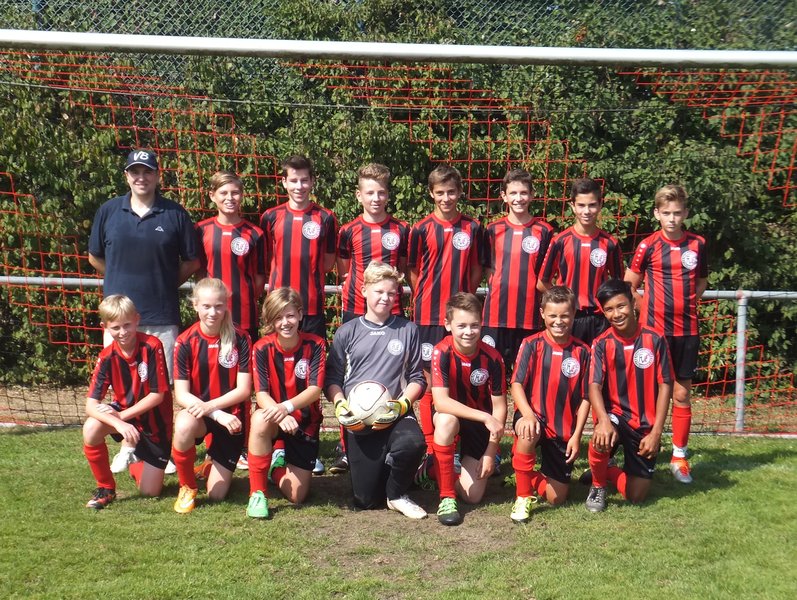 Eine Jugendfußballmannschaft posiert für ein Gruppenfoto in passenden rot-schwarz gestreiften Trikots. Sie stehen auf einem Rasen vor einem Tornetz, links steht ein Trainer. Ein Spieler hält einen Fußball. Im Hintergrund sind grüne Blätter und ein roter Zaun zu sehen.
