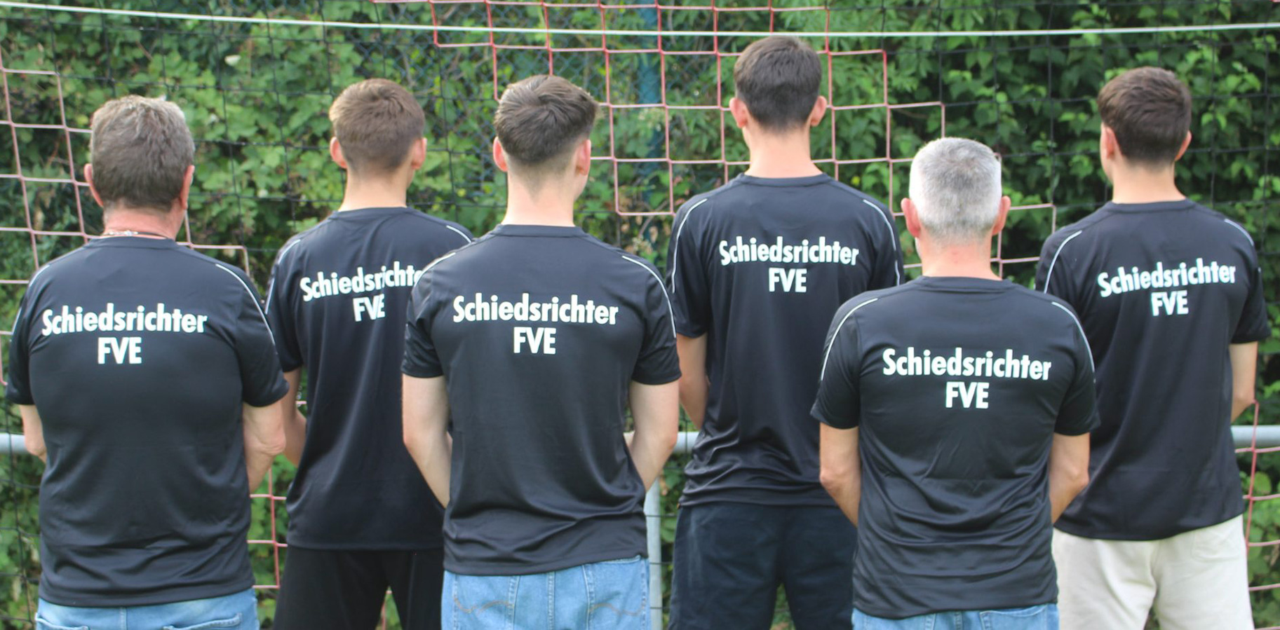 Sechs Personen stehen mit dem Rücken zur Kamera und tragen schwarze Shirts mit dem Text „Schiedsrichter FVE“. Sie stehen vor einem grünen Hintergrund und man sieht einen Teil eines Torpfostens.