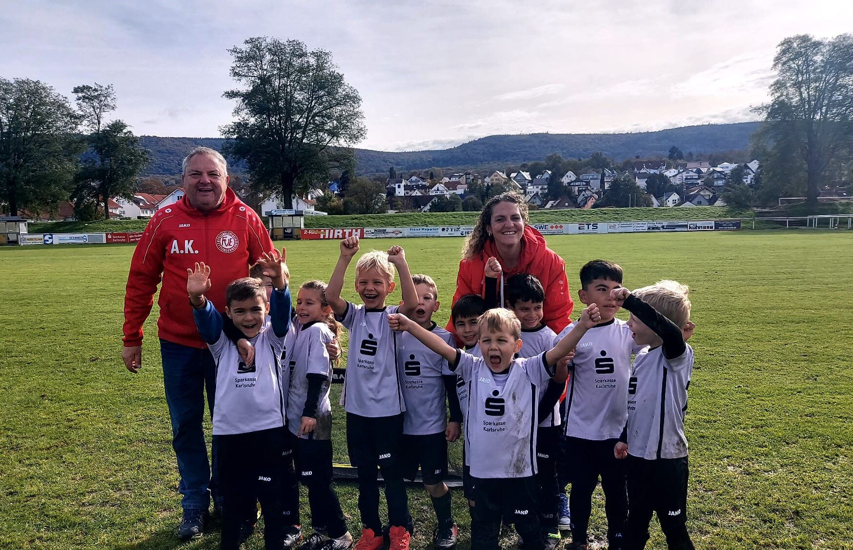 Eine Gruppe kleiner Kinder in weißen Fußballtrikots posiert fröhlich auf einer Wiese mit zwei Erwachsenen, von denen einer eine rote Jacke trägt. Sie lächeln und heben freudig die Arme. Im Hintergrund sind Bäume und eine Stadtlandschaft zu sehen.