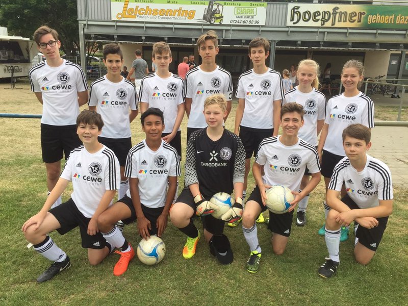 Eine Jugendfußballmannschaft posiert für ein Gruppenfoto auf einem Rasenplatz. Zwölf Spieler tragen schwarz-weiße Trikots, der Torwart trägt ein anderes Trikot. Im Hintergrund ist ein Gebäude mit Schildern zu sehen. Einige Spieler halten Fußbälle in den Händen.